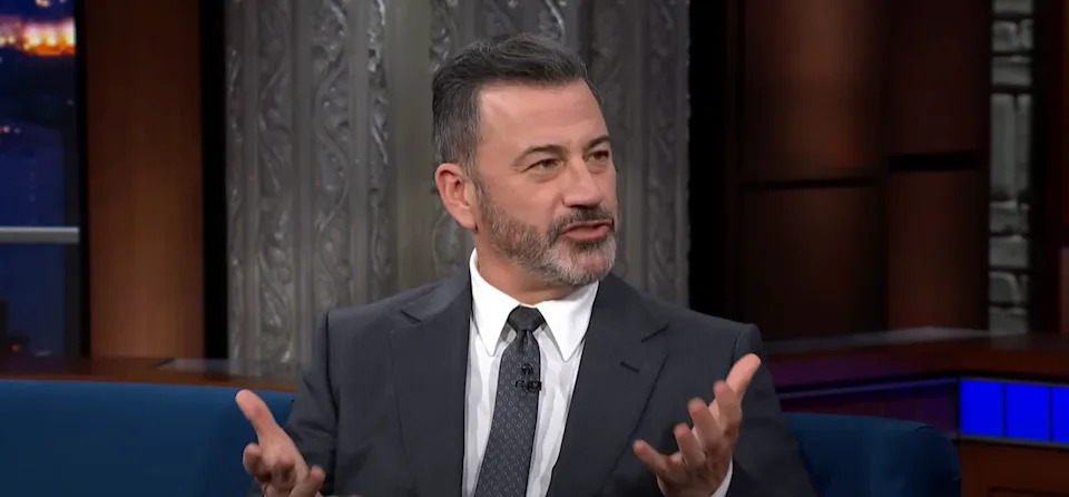 A man in a suit on a talk show set gestures animatedly during a conversation
