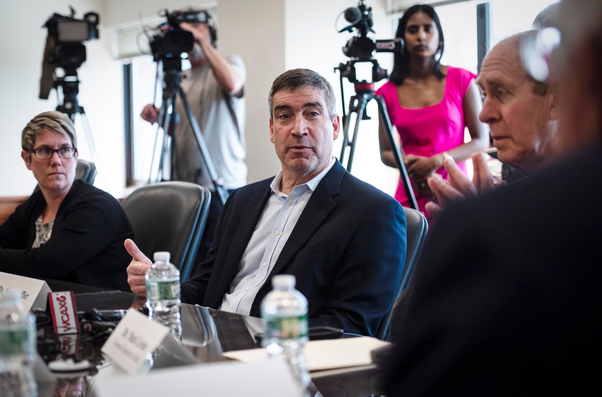 A man in a suit speaks during a meeting while seated at a conference table. Two people, one in a pink dress and another in a casual outfit, operate cameras in the background.