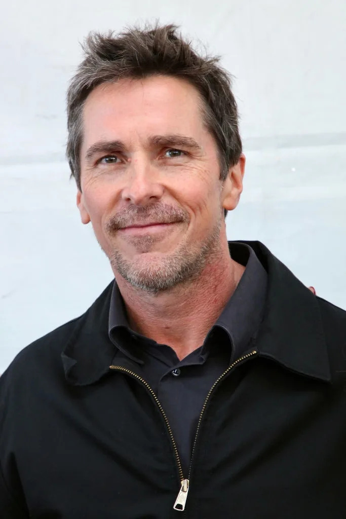 A person is wearing a casual, dark jacket over a collared shirt, smiling slightly, with short hair and a light beard