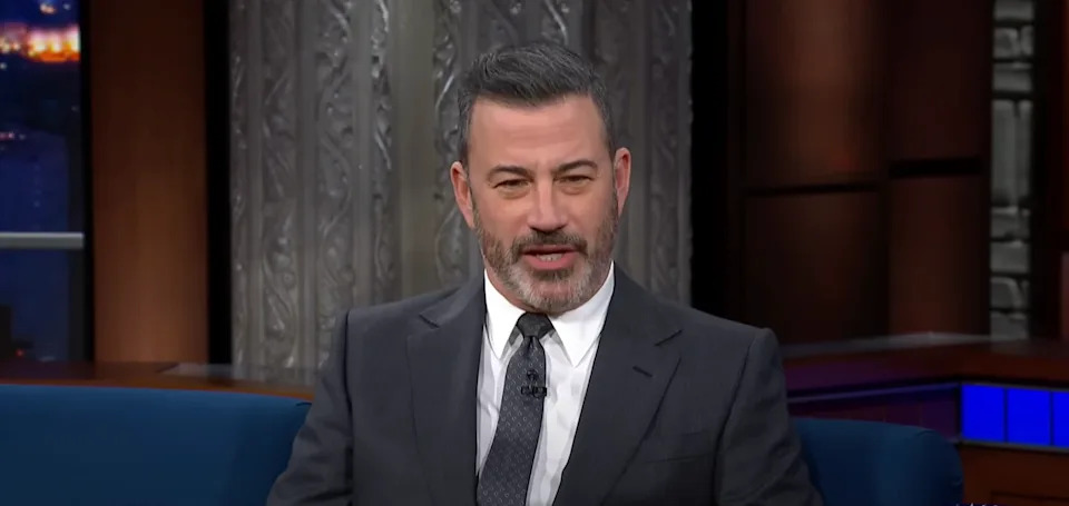 A man in a dark suit and patterned tie sits on a talk show set, speaking animatedly