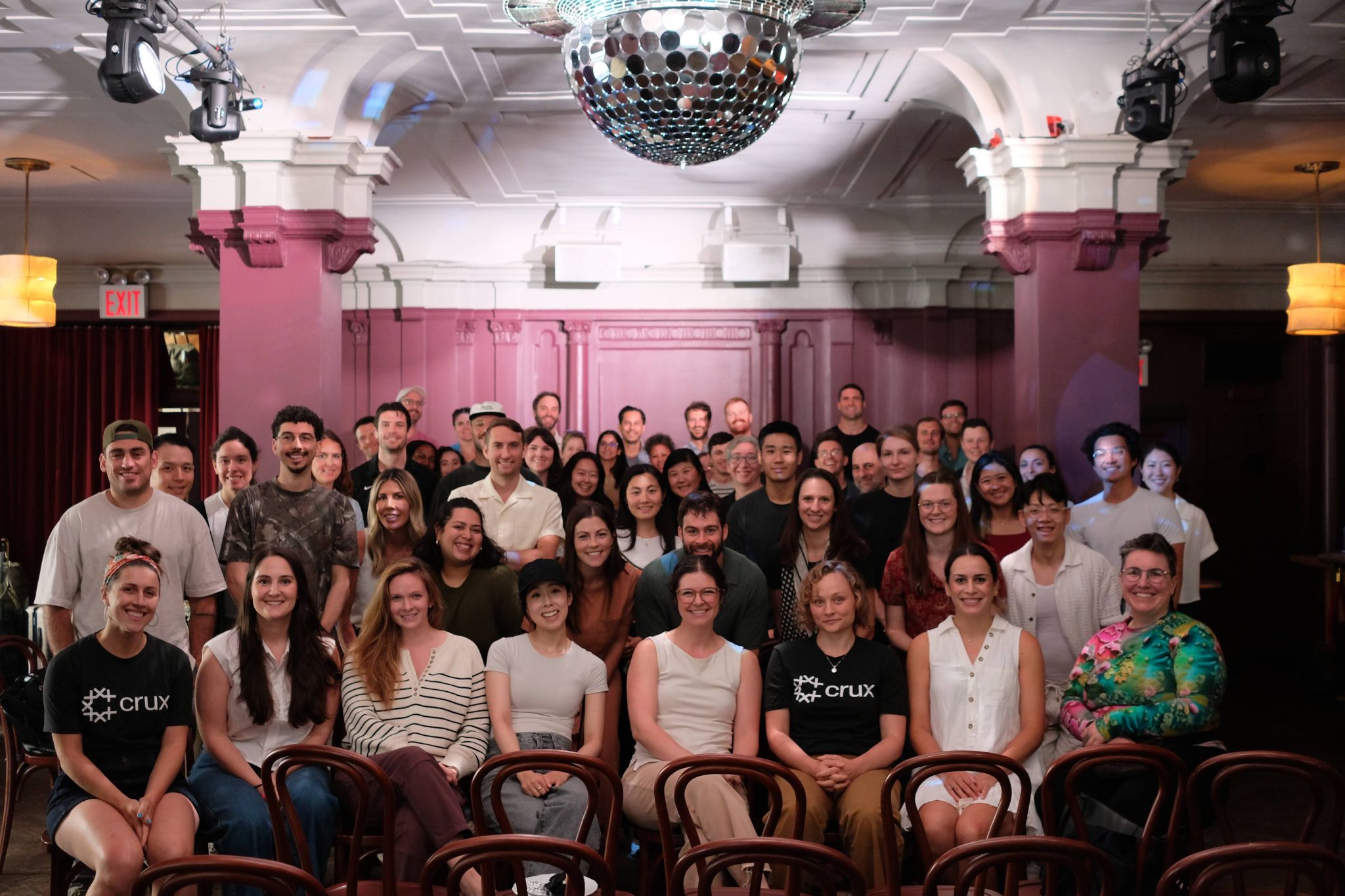 A group of people pose for a photo in a warmly lit room with pink walls and a disco ball overhead. Some in front wear shirts with a "crux" logo. Rows of empty chairs are visible.