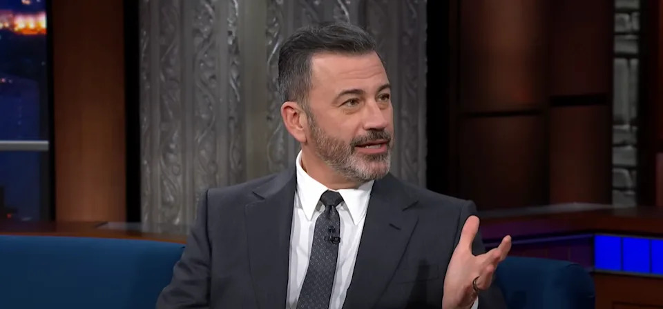 A man in a suit and tie is speaking animatedly while seated on a talk show set
