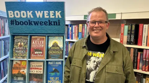 Brian McGilloway is in a green jacket and black top. 
He has glasses and has short brown hair. 
He is standing in a library with a number of books behind him. 
A stall with the words: 'Book Week #BookWeekNI' is visible beside him. 