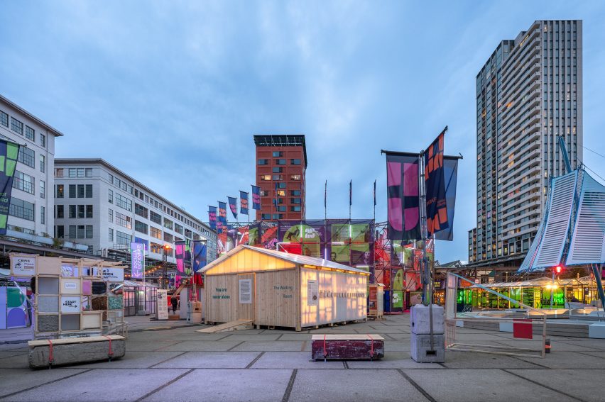 Photo of the Waiting Room pavilion lit up from within at dusk at Dutch Design Week 2025, surrounded by colourful flags and installations