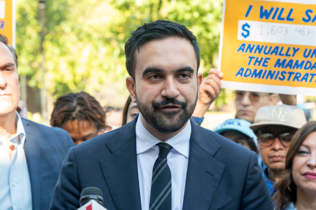 Zohran Mamdani, Democratic candidate for New York City mayor, speaking at a campaign event with a sign in the background displaying "I WILL SAVE $1,603.46 ANNUALLY UNDER THE MAMDANI ADMINISTRATION."