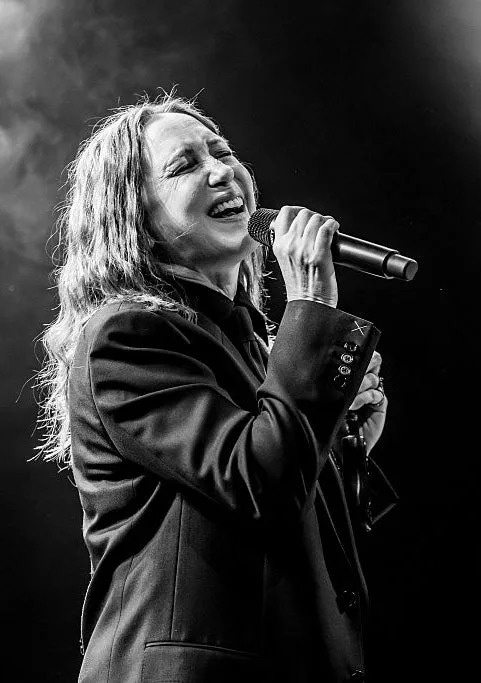 A person sings passionately on stage, holding a microphone, wearing a blazer. Another performer is visible in the background