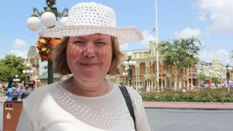 Liv Heeney A woman wearing a white sun hat and T-shirt stands outside on a sunny day. A building and trees can be seen behind her. People are crossing the road.