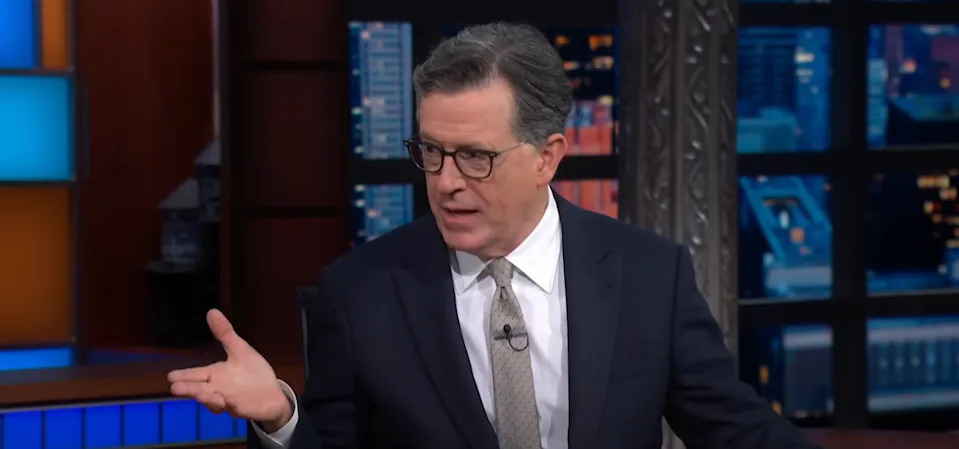 A person in a suit and tie is seated indoors, gesturing with one hand during a conversation on a talk show set