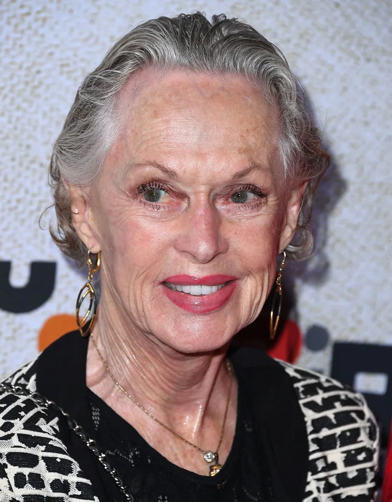 Woman smiling on a red carpet, wearing a patterned jacket and gold hoop earrings