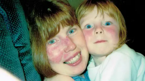 Liv Heeney A woman with blue eyes and red hair holds a young girl with blue eyes and red hair in her arms while sitting on a green sofa. The woman is wearing a blue top and the young girl is wearing a white T-shirt.