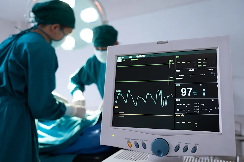 Medical professionals in surgical attire operate, with a monitor displaying vital signs in the foreground