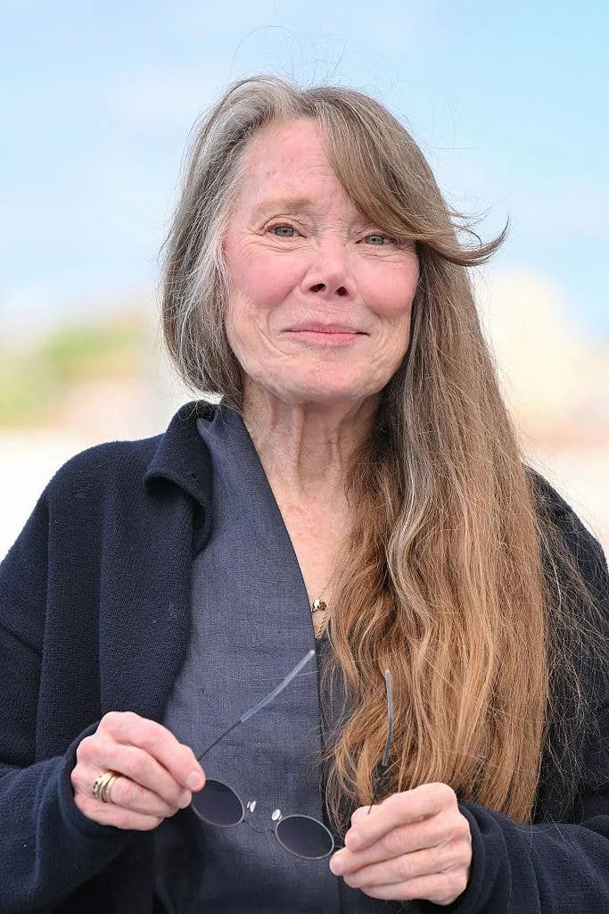 A person with long hair smiles while holding sunglasses. They wear a casual, dark outfit at an outdoor event