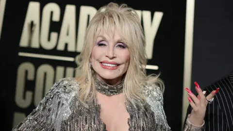 Getty Images Dolly Parton is wearing a silver dress. She has medium length blonde hair to her shoulders. She has long pink nails on her hand. She also has a silver necklace on. The background behind her is a billboard with white writing on a black  background.  