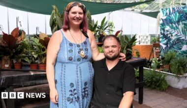A woman in a blue floral dress and headscarf stands beside a man seated in a wheelchair, wearing a black shirt. Both are inside a greenhouse filled with potted plants.