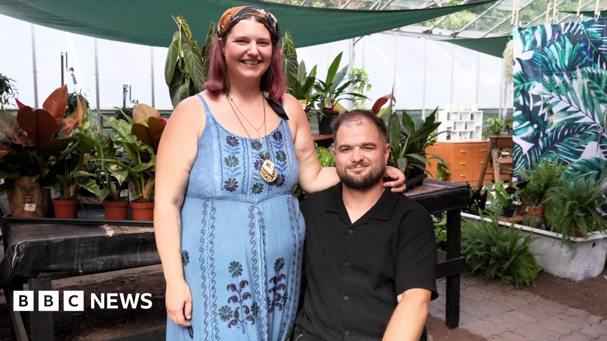 A woman in a blue floral dress and headscarf stands beside a man seated in a wheelchair, wearing a black shirt. Both are inside a greenhouse filled with potted plants.