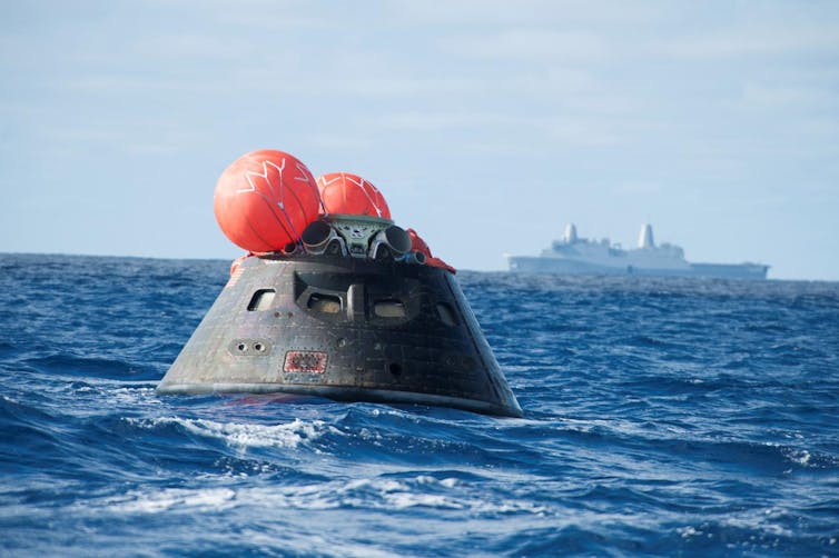A capsule, with buoys attached, floating through the ocean with a large vessel in the background.