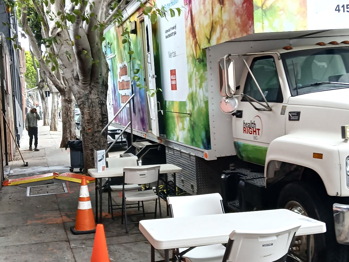 A mobile health clinic truck is parked on a city street beside folding tables and chairs, with orange traffic cones placed nearby.