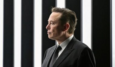Elon Musk in profile wearing a suit and tie, standing in front of vertical black and white striped panels.