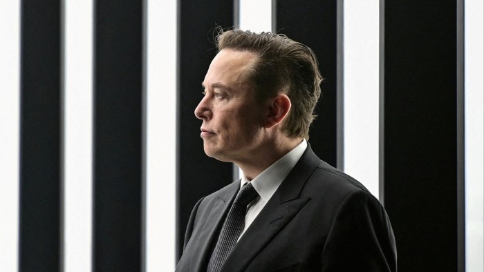 Elon Musk in profile wearing a suit and tie, standing in front of vertical black and white striped panels.