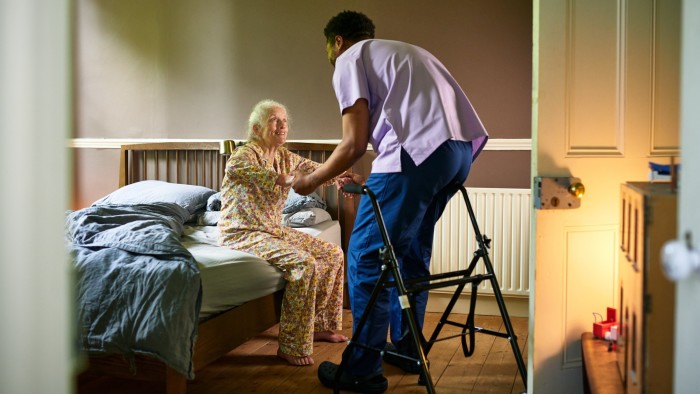 Senior woman in floral pajamas smiling and holding hands with a male carer, preparing to stand up from her bed.