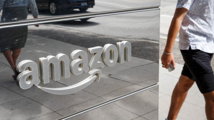 The Amazon logo on a reflective office wall, with pedestrians and street activity visible in the reflection.