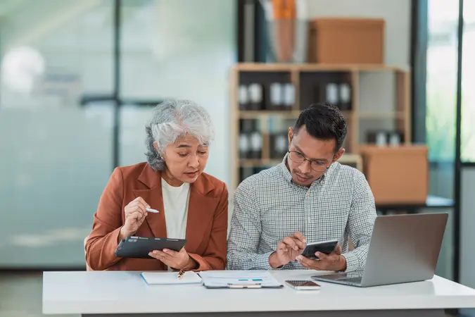 Senior Asian business woman and young man working together analyzing finances senior staff teach marketing and planning to younger staff woman holding graph and chart with a happy smile.
