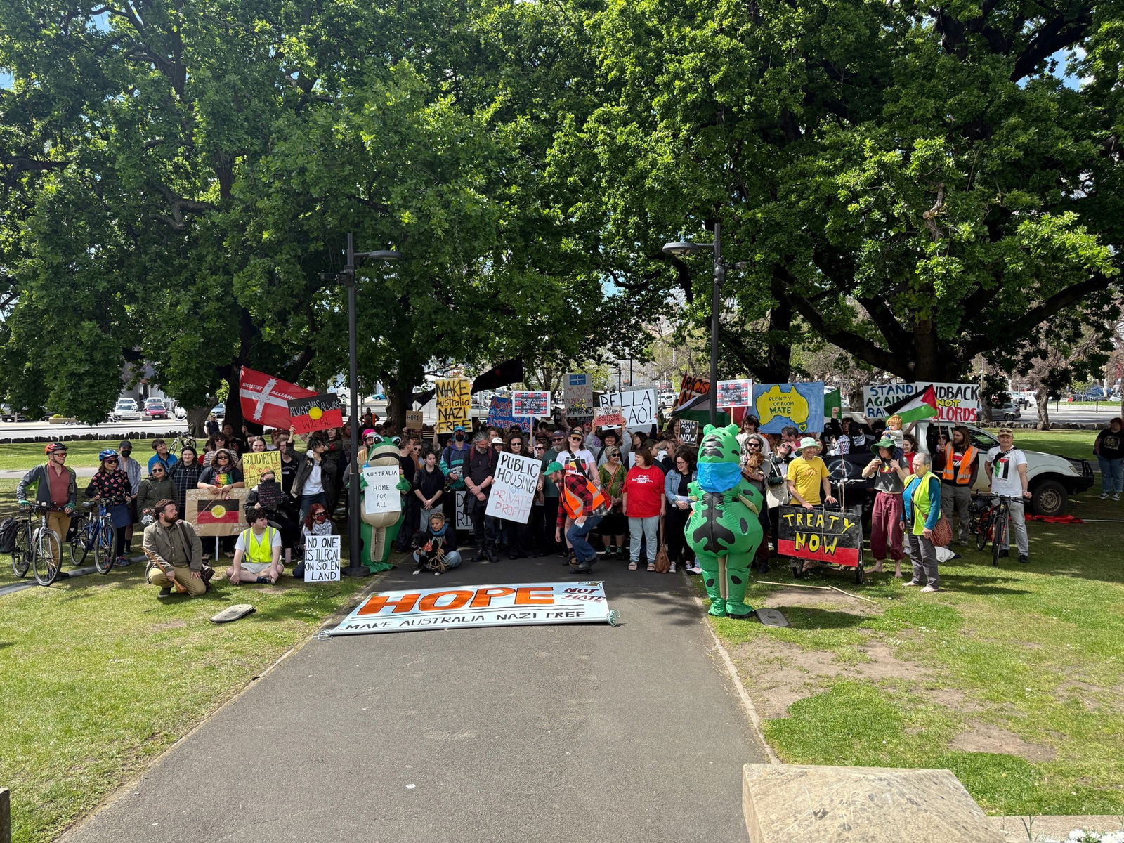 A crowd of people holding signs and Aboriginal and Palestinian flags stand in a park