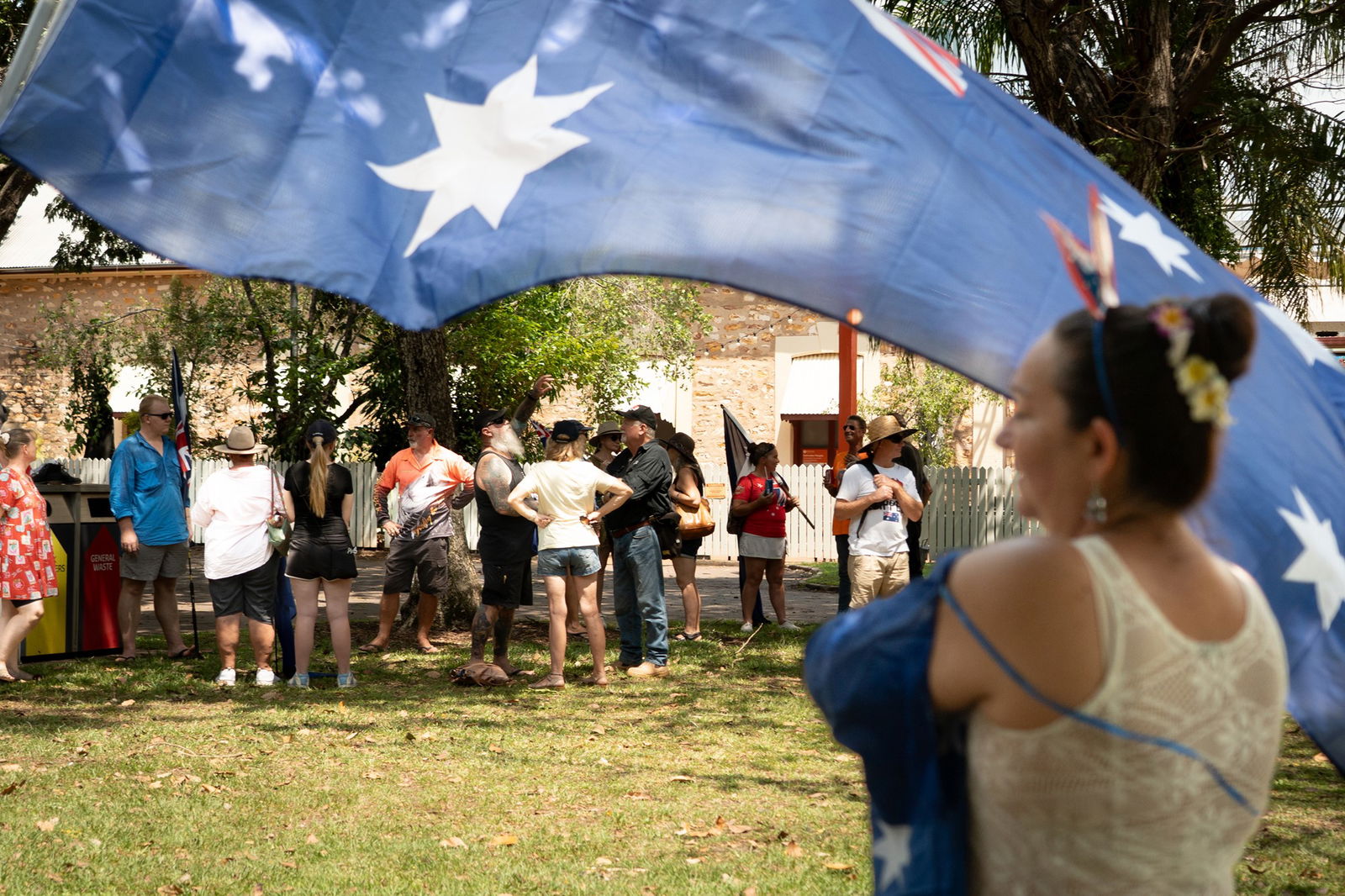 A crowd of people, mostly in shorts and tshirts standing in a park, Australian flag flying in foreground along with woman out of focus in front of it.