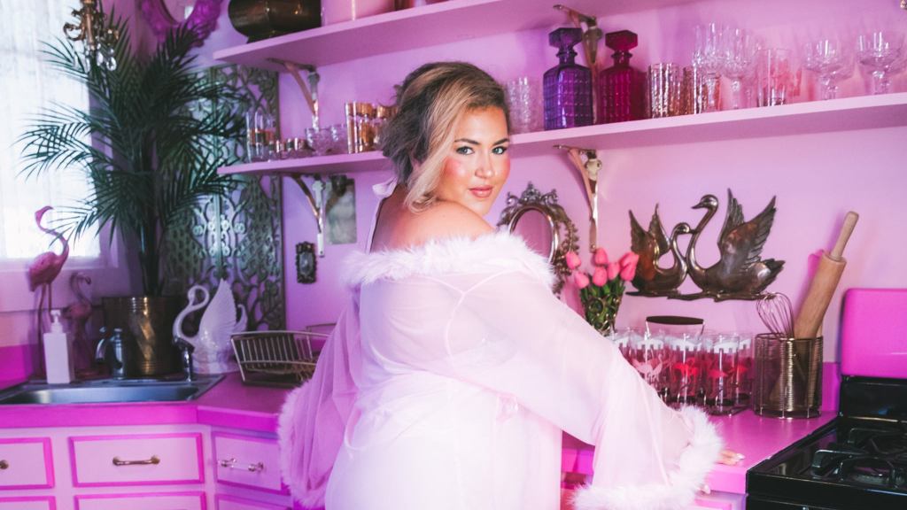 A blonde woman in a pink robe looks over her shoulder in a pink kitchen.