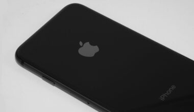 A close-up of a black iPhone placed on a white surface, showing the Apple logo and part of the word "iPhone" on the back.