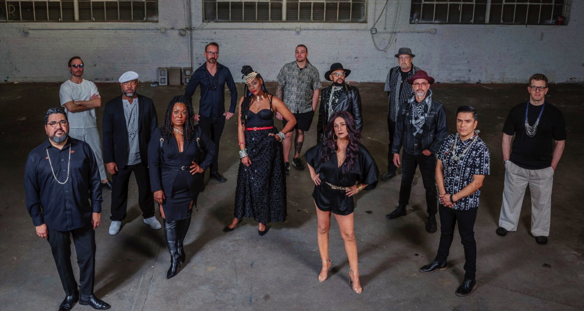 A group of 13 musicians standing in front of a gray wall.