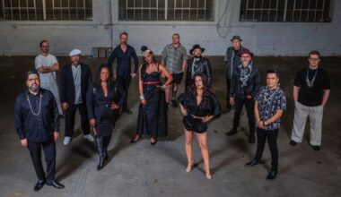 A group of 13 musicians standing in front of a gray wall.