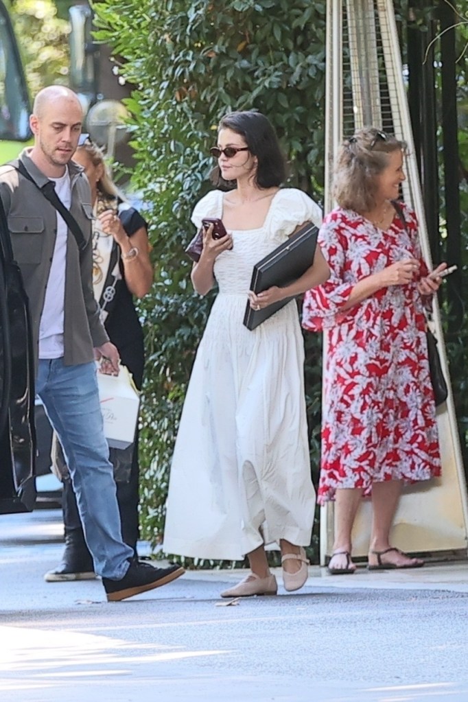 Selena Gomez in a white dress, holding a phone and a black box, with a bodyguard and another woman.