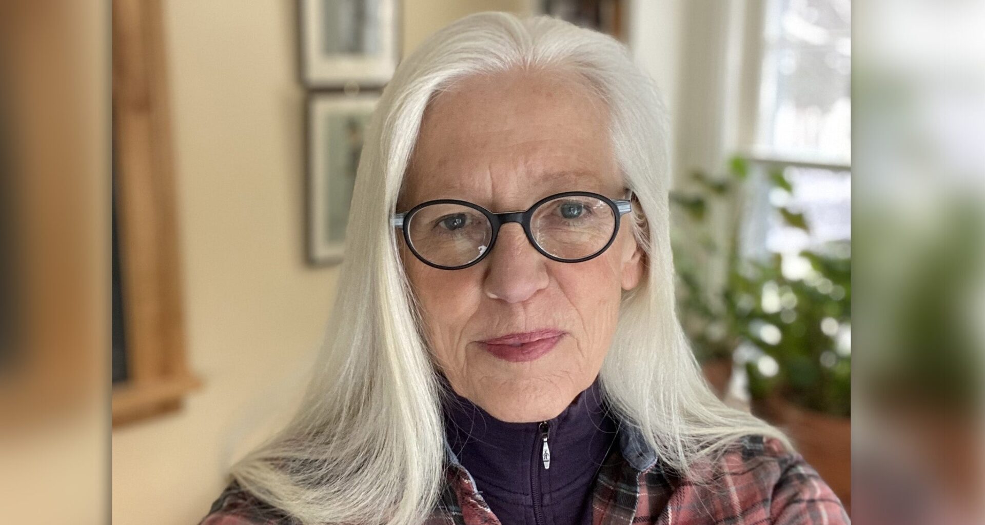 An older woman with long white hair and glasses looks at the camera, indoors, with plants and framed pictures visible in the background.