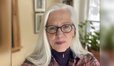 An older woman with long white hair and glasses looks at the camera, indoors, with plants and framed pictures visible in the background.