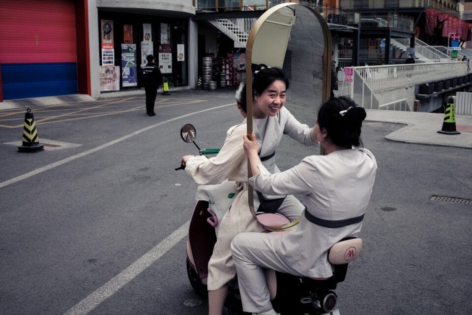 a woman riding on the back of a moped holds a mirror, revealing a smiling reflection of herself