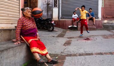 three children in the street watch as their basketball is about to hit an old woman in the face