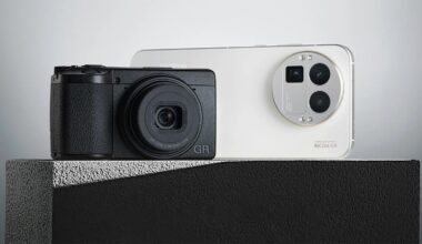 A black compact camera and a white smartphone with triple rear cameras are displayed side by side on a textured gray surface against a minimalist background.