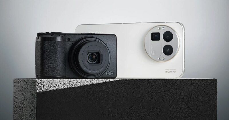 A black compact camera and a white smartphone with triple rear cameras are displayed side by side on a textured gray surface against a minimalist background.