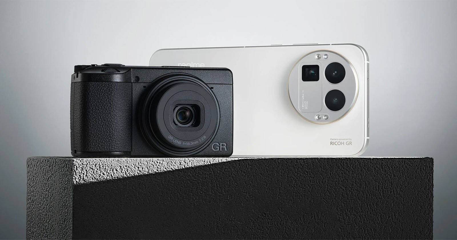 A black compact camera and a white smartphone with triple rear cameras are displayed side by side on a textured gray surface against a minimalist background.