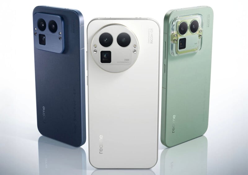 Three Realme smartphones in blue, white, and green are displayed standing upright. Each phone features a large, square camera module with prominent circular lenses on the back. The background is minimal and light-colored.