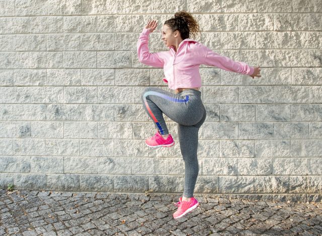 Joyful sporty Latin girl jumping high and raising knee. Street dancer warming up intensively before street workout. Student enjoying free fitness. Fitness and street workout concept