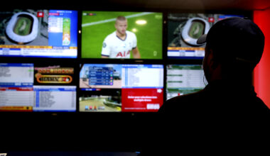 A man shrouded in shadow watches sports and engages in sports betting on a large screen.