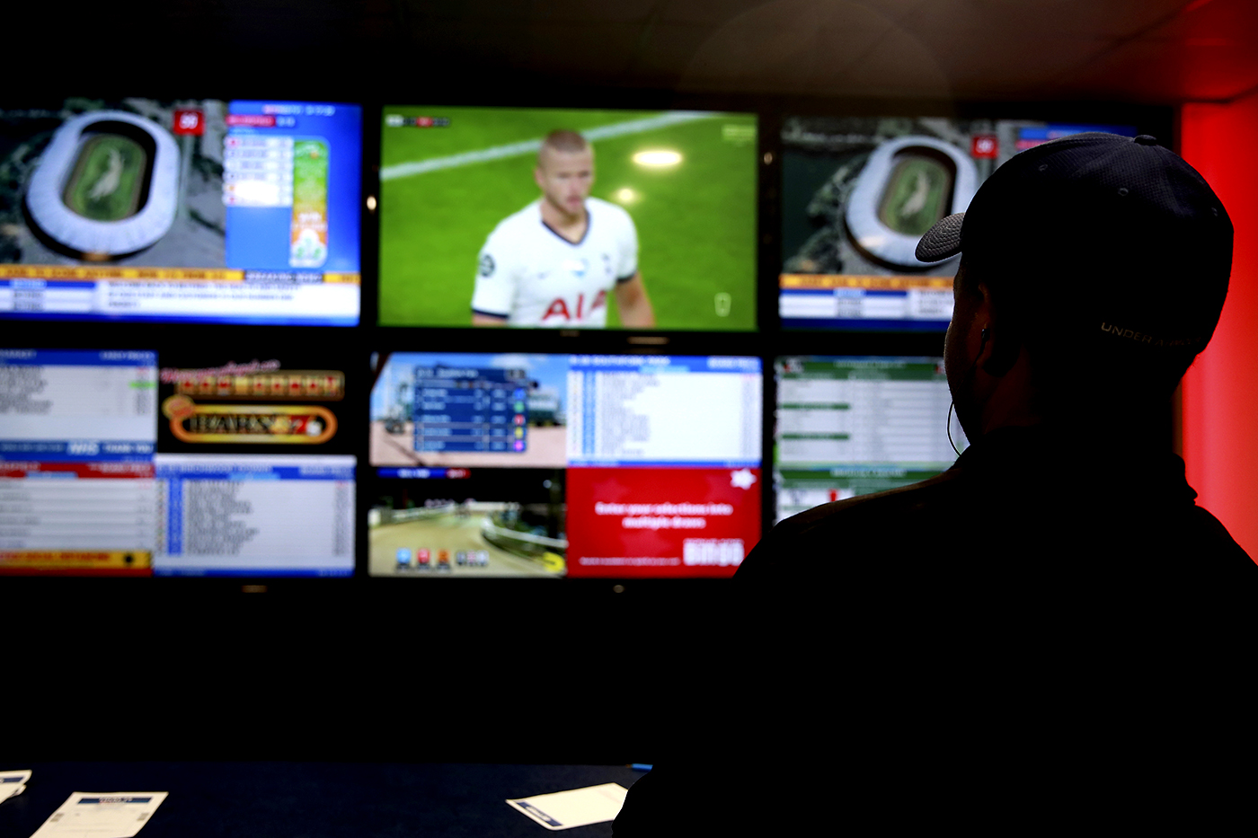 A man shrouded in shadow watches sports and engages in sports betting on a large screen.