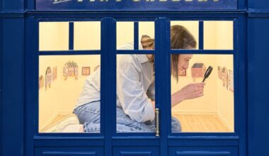 a person kneels inside of a 1/6-scale gallery, holding a magnifying glass up to a painting