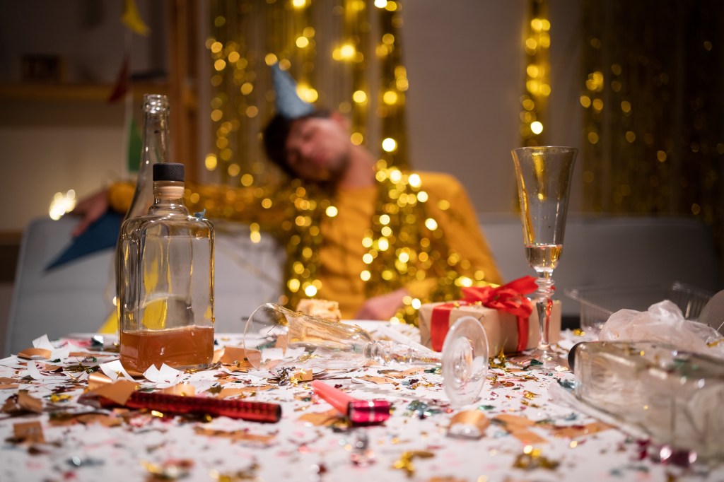 A man in a party hat passed out on a sofa after a party, with a messy table of bottles, confetti, and party debris in the foreground.