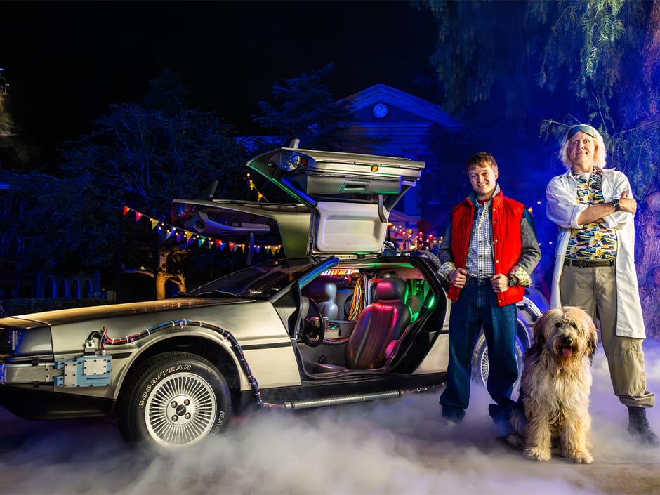 Two people and a dog stand by a car with gull-wing doors open, reminiscent of "Back to the Future." The misty scene, set at Universal Fan Fest Nights, is aglow with colorful lights and trees in the background.
