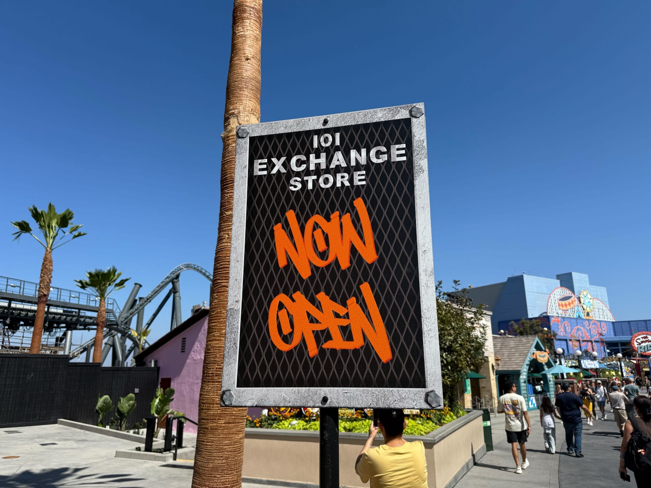 A sign with "101 Exchange Store NOW OPEN" stands outside under a clear blue sky, near Universal Studios—not a Disney park castle.
