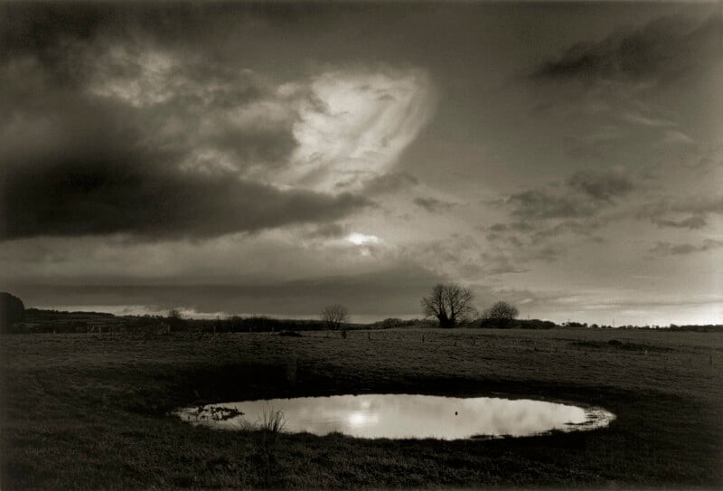 A small, circular pond reflects a dramatic, cloudy sky in a grassy field. Bare trees and distant hills are visible under the moody, low light, creating a tranquil and slightly somber rural landscape.