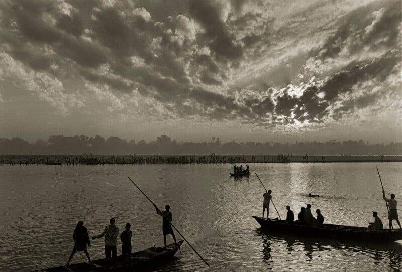 Boats with standing and seated people are silhouetted against a dramatic cloudy sky at sunrise or sunset over a calm river, creating a peaceful and scenic atmosphere.